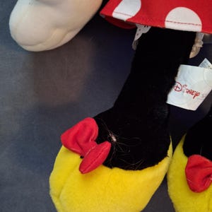 May include: Close-up of a Minnie Mouse plush toy's foot. The foot is yellow with a red bow and black leg. The red dress with white polka dots is visible. A Disney tag is attached.