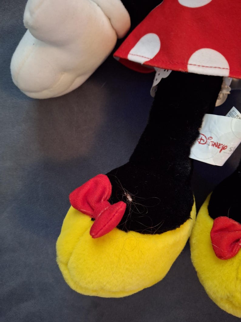 May include: Close-up of a Minnie Mouse plush toy's foot. The foot is yellow with a red bow and black leg. The red dress with white polka dots is visible. A Disney tag is attached.