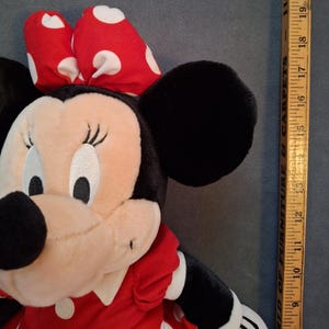 May include: A plush Minnie Mouse doll with a red dress and a red bow with white polka dots. The doll has black ears and a beige face. A wooden ruler is next to the doll, measuring approximately 48 cm.