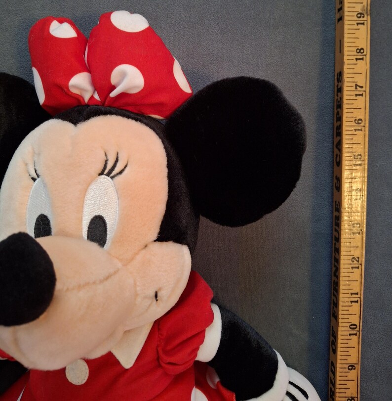 May include: A plush Minnie Mouse doll with a red dress and a red bow with white polka dots. The doll has black ears and a beige face. A wooden ruler is next to the doll, measuring approximately 48 cm.