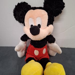 May include: A plush Mickey Mouse toy. The toy features a black head and ears, a tan face, white gloves, a red shirt with white buttons, black shorts, and large yellow shoes. The toy is sitting upright.