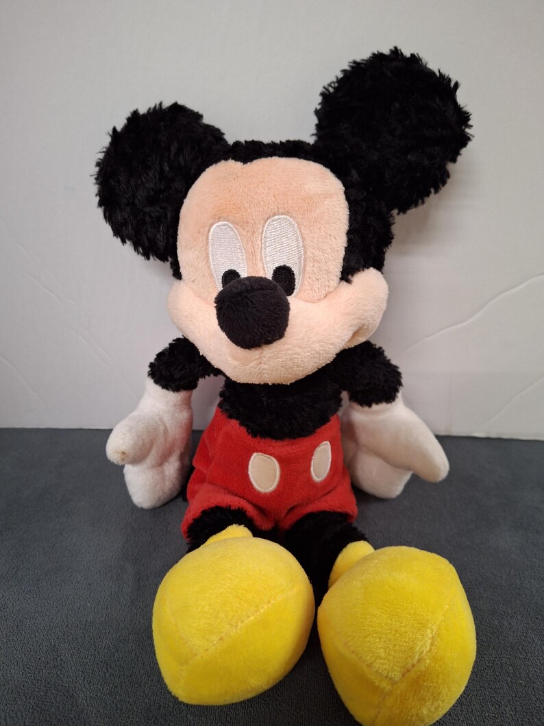 May include: A plush Mickey Mouse toy. The toy features a black head and ears, a tan face, white gloves, a red shirt with white buttons, black shorts, and large yellow shoes. The toy is sitting upright.
