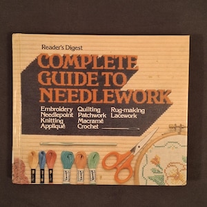 May include: A book titled "Complete Guide to Needlework" by Reader's Digest. The cover features orange text on a light brown background, with a list of needlework techniques. Embroidery floss, scissors, and a needlepoint hoop are also visible.