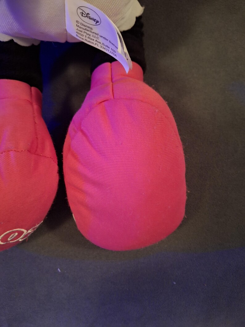 May include: Close-up of a plush toy's bright pink foot. The foot is made of soft fabric and has a rounded shape. A white tag with the Disney logo is visible. The toy is likely a character from Disney.