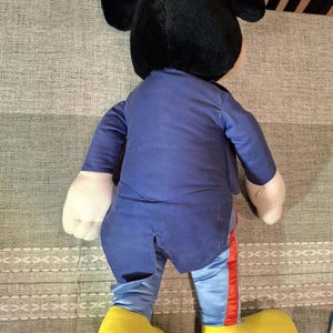 May include: A large Mickey Mouse plush toy, viewed from the back. The plush has a black head with large ears, a blue outfit, and yellow shoes. The pants have a red stripe down the side. The toy is on a patterned surface.