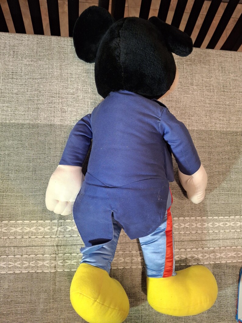 May include: A large Mickey Mouse plush toy, viewed from the back. The plush has a black head with large ears, a blue outfit, and yellow shoes. The pants have a red stripe down the side. The toy is on a patterned surface.