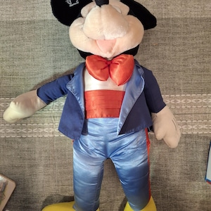 May include: A large Mickey Mouse plush toy wearing a blue tuxedo jacket, pants, and a red bow tie. The plush has black ears, a white face, and yellow shoes. The toy is approximately 30 inches tall.