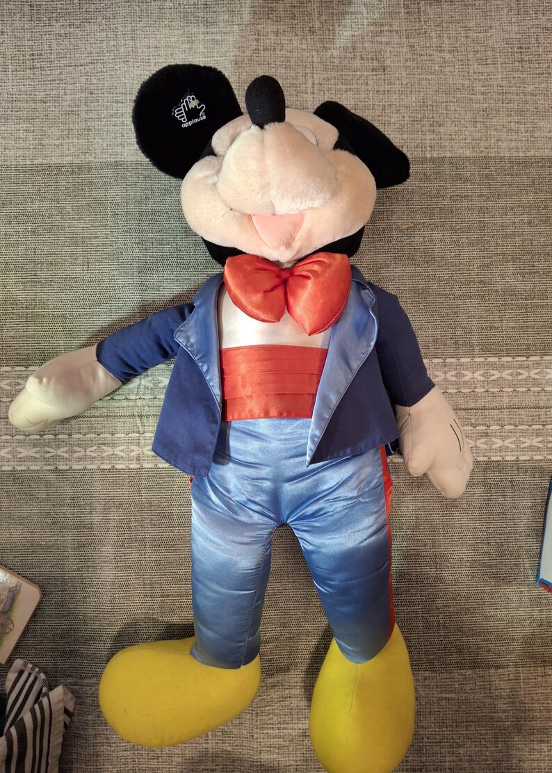 May include: A large Mickey Mouse plush toy wearing a blue tuxedo jacket, pants, and a red bow tie. The plush has black ears, a white face, and yellow shoes. The toy is approximately 30 inches tall.