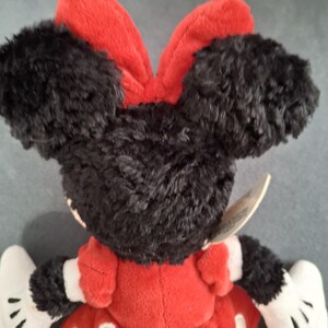 May include: A plush Minnie Mouse doll, viewed from the back. The doll features black fur ears and head, a red bow, and a red dress with white polka dots. White gloves and a tag are also visible.