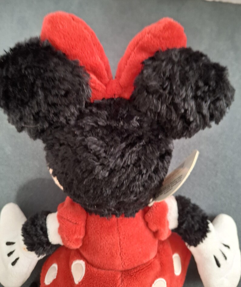 May include: A plush Minnie Mouse doll, viewed from the back. The doll features black fur ears and head, a red bow, and a red dress with white polka dots. White gloves and a tag are also visible.