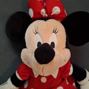 May include: A Minnie Mouse soft toy. The toy features a cream-coloured face, black ears, and a red dress with white polka dots. A matching red bow with white polka dots is on her head. The toy's arms and hands are white.
