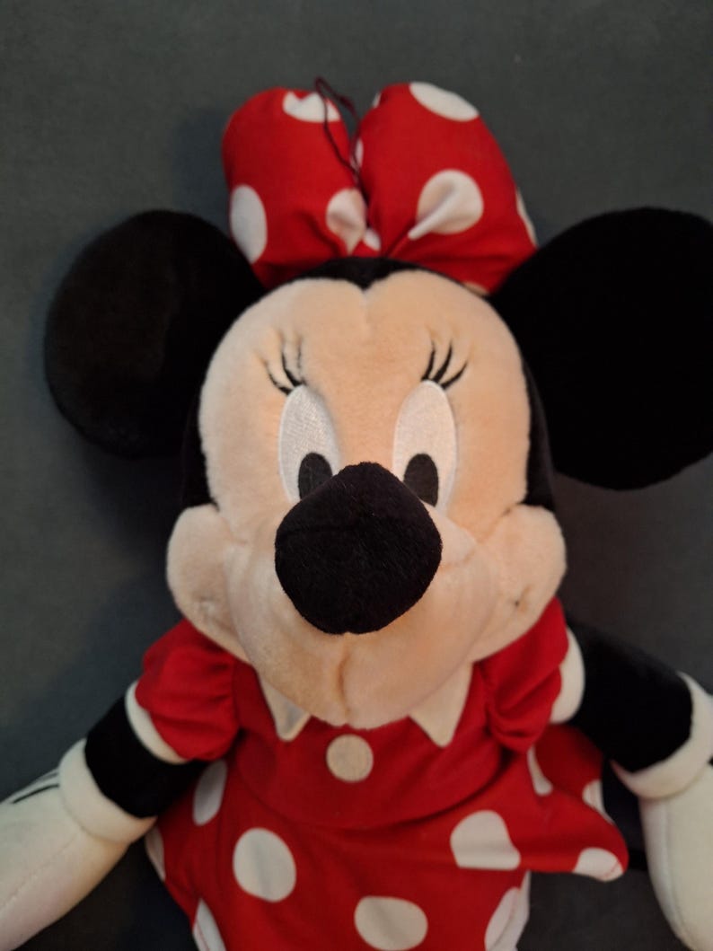 May include: A Minnie Mouse soft toy. The toy features a cream-coloured face, black ears, and a red dress with white polka dots. A matching red bow with white polka dots is on her head. The toy's arms and hands are white.