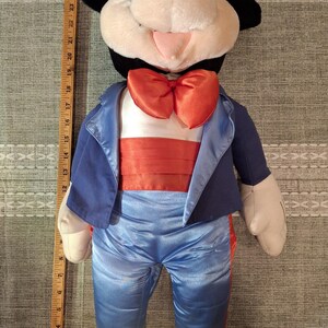 May include: A plush Mickey Mouse doll wearing a blue tuxedo jacket, pants, and an orange bow tie. The doll has black ears, a white face, and yellow shoes. The doll is approximately 28 inches tall.