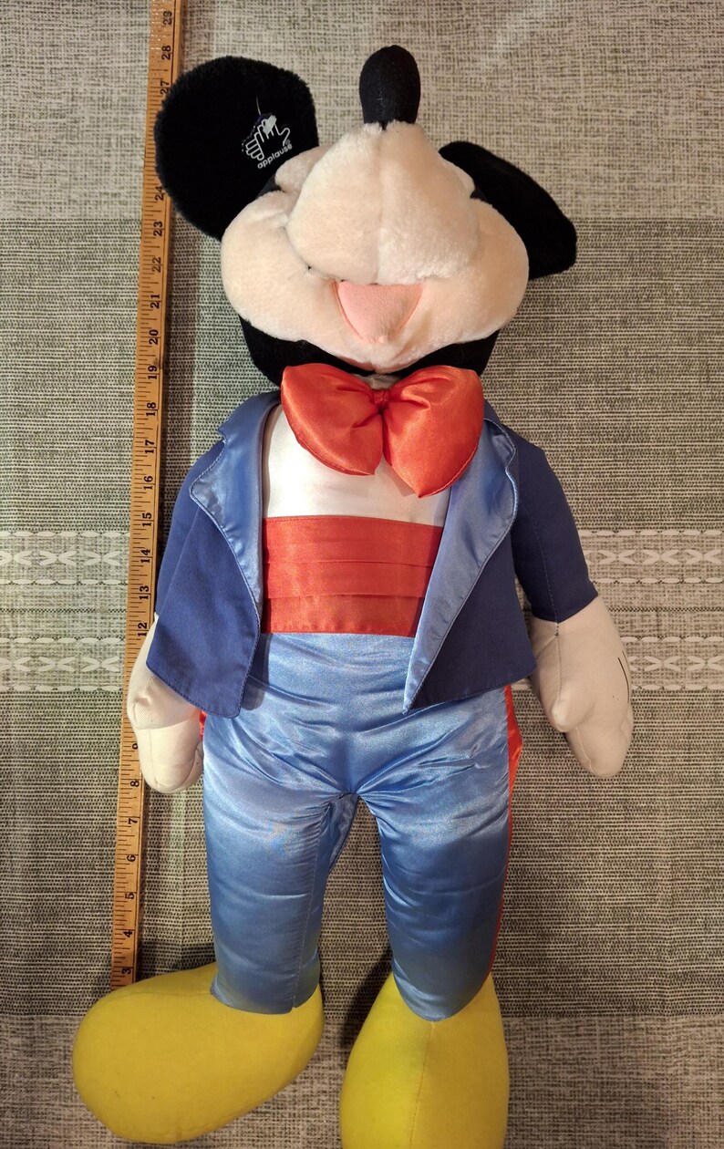 May include: A plush Mickey Mouse doll wearing a blue tuxedo jacket, pants, and an orange bow tie. The doll has black ears, a white face, and yellow shoes. The doll is approximately 28 inches tall.