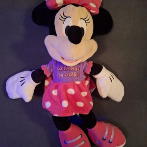 May include: A plush Minnie Mouse doll, featuring a pink polka-dot dress, matching bow, and red shoes. The doll has black ears, a black nose, and white gloves. The text "MINNIE MOUSE" is printed on the purple shirt.