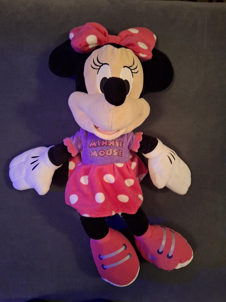 May include: A plush Minnie Mouse doll, featuring a pink polka-dot dress, matching bow, and red shoes. The doll has black ears, a black nose, and white gloves. The text "MINNIE MOUSE" is printed on the purple shirt.