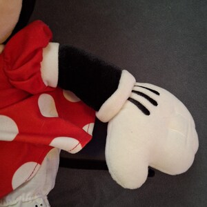 May include: A plush toy featuring a red dress with white polka dots and a white trim. The toy has a black arm with a white cuff and a white glove with black lines. The background is a solid grey.