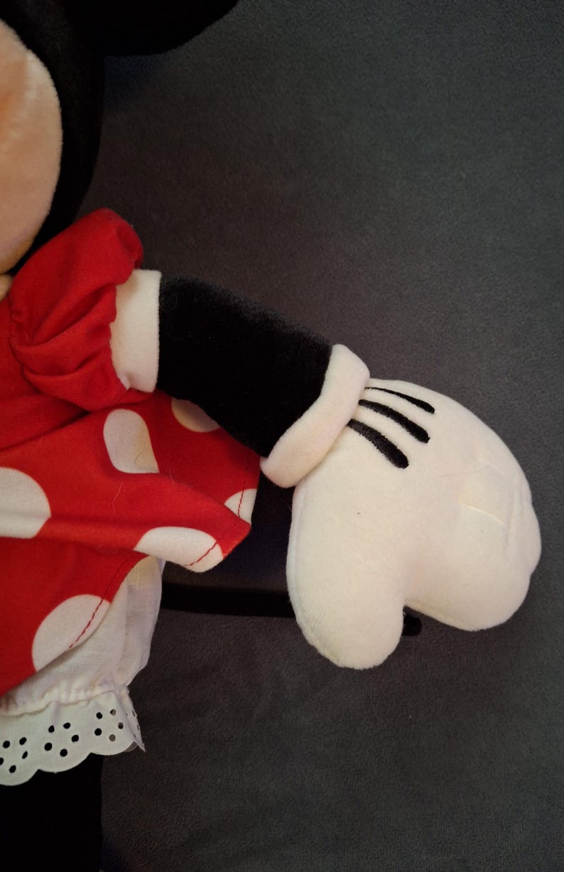 May include: A plush toy featuring a red dress with white polka dots and a white trim. The toy has a black arm with a white cuff and a white glove with black lines. The background is a solid grey.