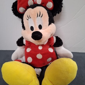May include: A plush Minnie Mouse doll. The doll has a black head with large, round eyes, a black nose, and a red bow with white polka dots. She wears a red dress with white polka dots and yellow shoes.