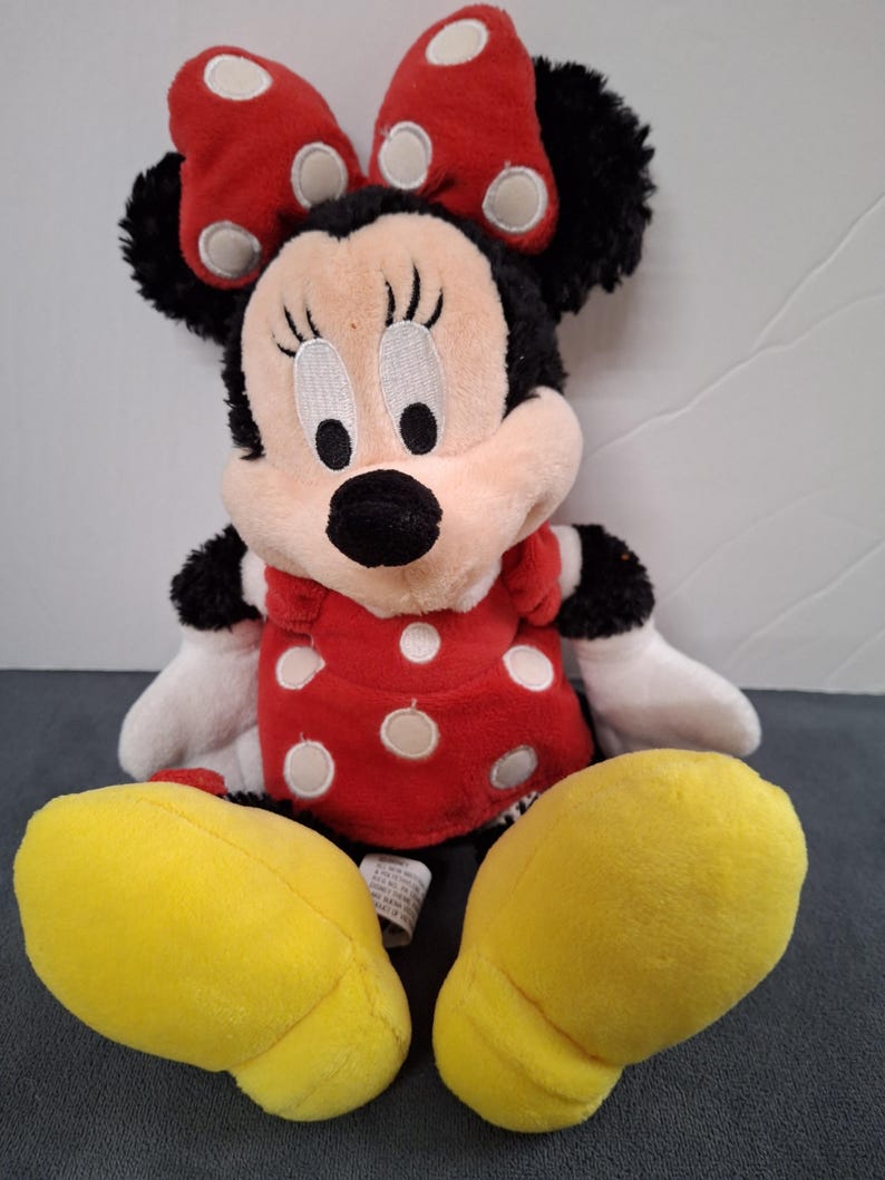 May include: A plush Minnie Mouse doll. The doll has a black head with large, round eyes, a black nose, and a red bow with white polka dots. She wears a red dress with white polka dots and yellow shoes.
