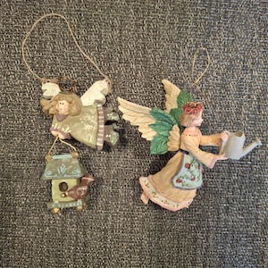 May include: Two angel ornaments with wings and hanging loops. One angel holds a birdhouse, the other holds a watering can. The ornaments are painted in pastel colors with floral and nature-inspired details.