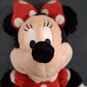May include: A plush Minnie Mouse doll. The doll has a red dress with white polka dots, a matching bow, and black ears. The face is beige with black eyes, eyelashes, and a black nose. The doll's arms are black and white.