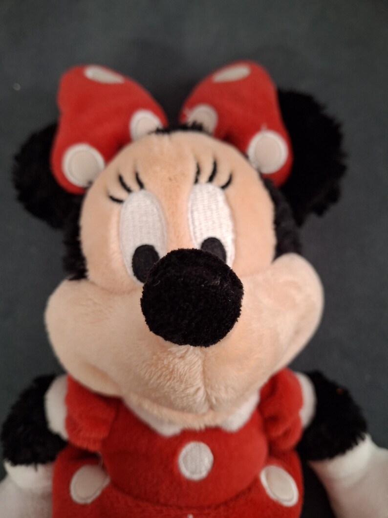 May include: A plush Minnie Mouse doll. The doll has a red dress with white polka dots, a matching bow, and black ears. The face is beige with black eyes, eyelashes, and a black nose. The doll's arms are black and white.