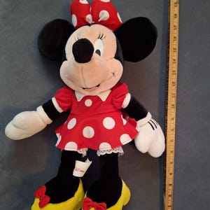 May include: A plush Minnie Mouse doll, wearing a red dress with white polka dots, a matching bow, and yellow shoes. The doll has black ears, arms, and legs, with white gloves. A wooden ruler is next to the doll, measuring up to 53 cm.