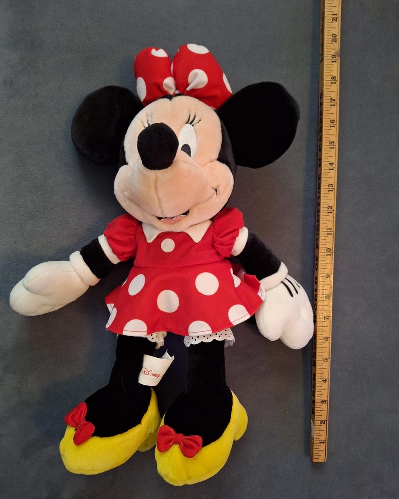 May include: A plush Minnie Mouse doll, wearing a red dress with white polka dots, a matching bow, and yellow shoes. The doll has black ears, arms, and legs, with white gloves. A wooden ruler is next to the doll, measuring up to 53 cm.