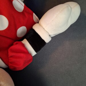 May include: A plush toy arm and hand, with a red sleeve featuring white polka dots and a white glove. The arm has a black cuff with white trim. The background is a solid grey.