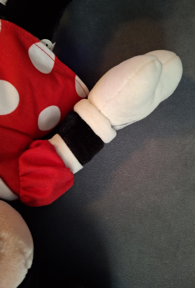 May include: A plush toy arm and hand, with a red sleeve featuring white polka dots and a white glove. The arm has a black cuff with white trim. The background is a solid grey.
