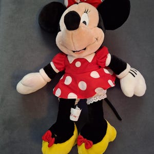 May include: A Minnie Mouse plush doll. The doll has a red dress with white polka dots, a matching bow, and yellow shoes. The doll has black ears, black legs, and white gloves. A Disney tag is attached to the doll.