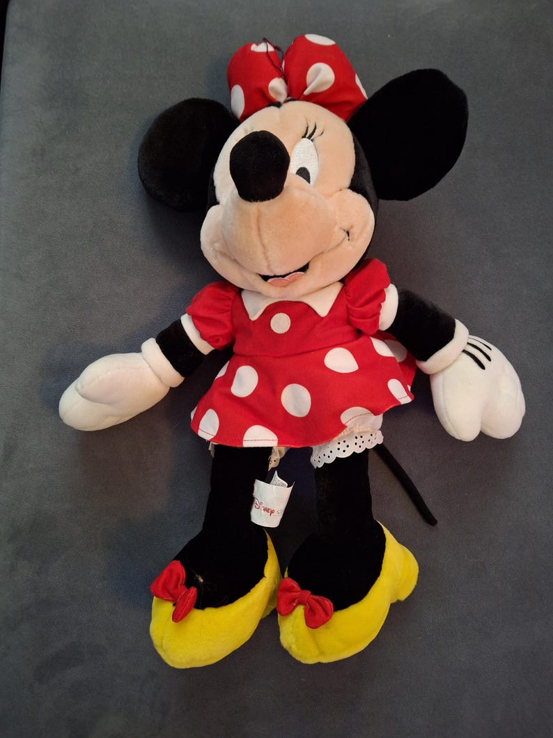 May include: A Minnie Mouse plush doll. The doll has a red dress with white polka dots, a matching bow, and yellow shoes. The doll has black ears, black legs, and white gloves. A Disney tag is attached to the doll.