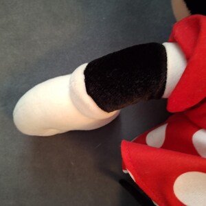 May include: Close-up of a plush toy arm and dress. The arm is white with a black sleeve and the dress is red with white polka dots. The toy is on a grey surface.