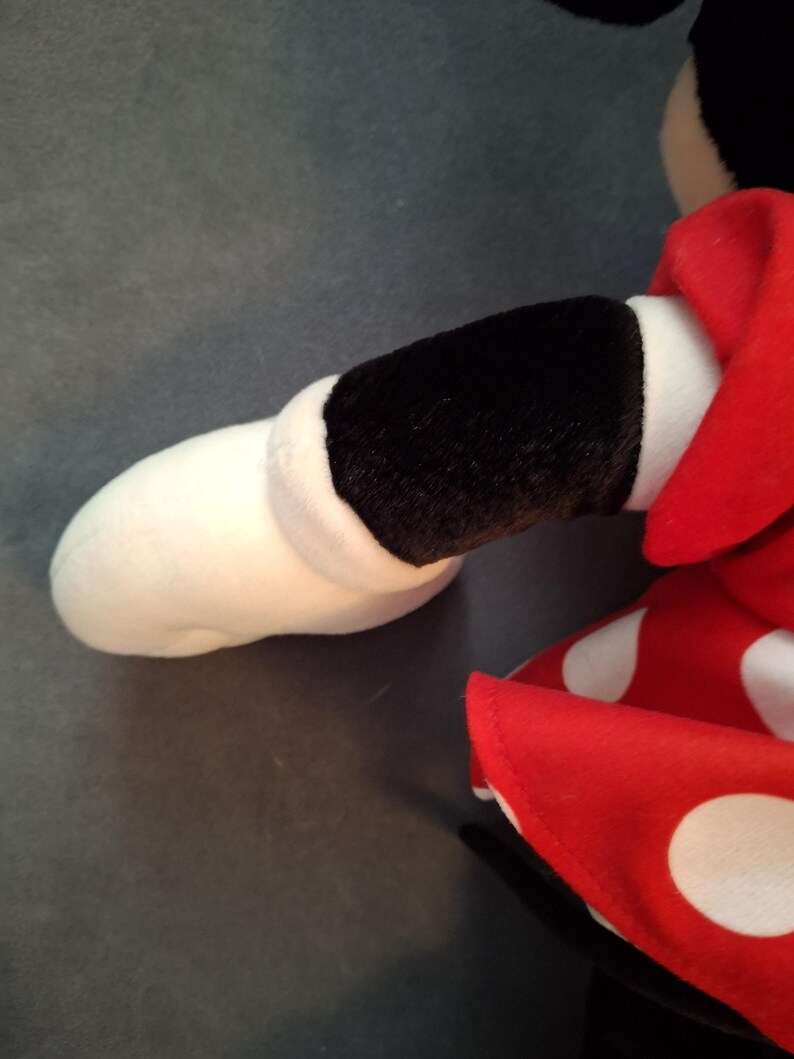 May include: Close-up of a plush toy arm and dress. The arm is white with a black sleeve and the dress is red with white polka dots. The toy is on a grey surface.