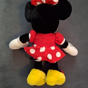 May include: A Minnie Mouse soft toy, seen from behind, with a red dress and white polka dots, a matching bow, and yellow shoes. The doll has black ears, white gloves, and a black face.