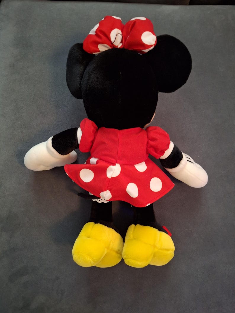 May include: A Minnie Mouse soft toy, seen from behind, with a red dress and white polka dots, a matching bow, and yellow shoes. The doll has black ears, white gloves, and a black face.