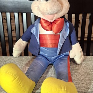May include: A large plush Mickey Mouse doll wearing a blue tuxedo jacket, pants, and a red bow tie and cummerbund. The doll has large yellow shoes and black ears. The doll is sitting on a gray patterned surface.
