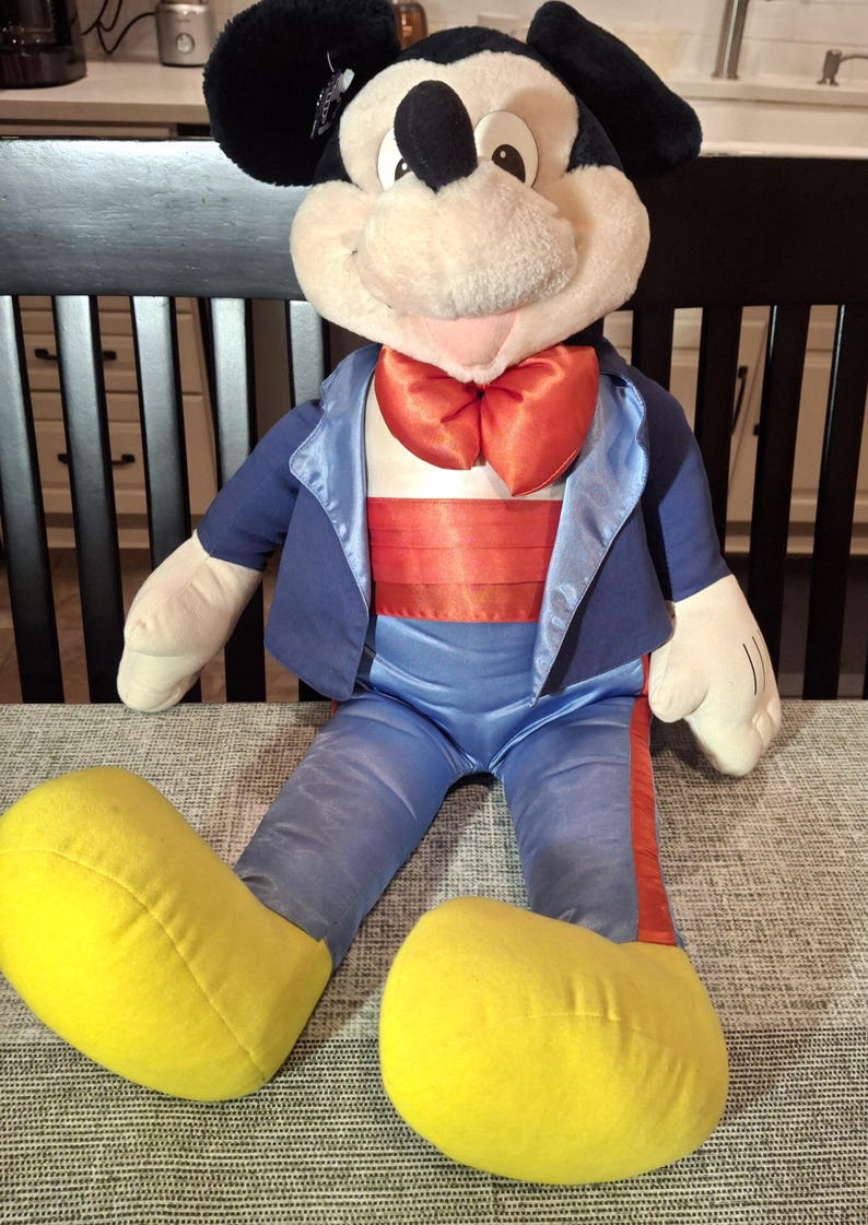 May include: A large plush Mickey Mouse doll wearing a blue tuxedo jacket, pants, and a red bow tie and cummerbund. The doll has large yellow shoes and black ears. The doll is sitting on a gray patterned surface.