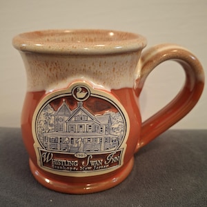 May include: A ceramic mug with a brown and cream speckled finish. The mug features a detailed illustration of a building with the text "Whistling Swan Inn, Stanhope, New Jersey" and the year "1905". The handle and body are a reddish-brown color.