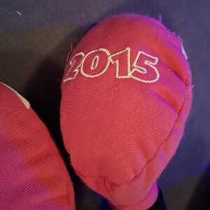 May include: Close-up of a red fabric item with the year "2015" embroidered in white. The fabric appears to be a soft material, possibly cotton or a similar textile. The item is partially visible against a dark background.