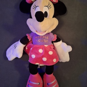 May include: A plush Minnie Mouse doll with a pink polka dot bow, dress, and shoes. The doll has black ears, a black nose, and a purple shirt with the words "Minnie Mouse". The doll has white gloves.