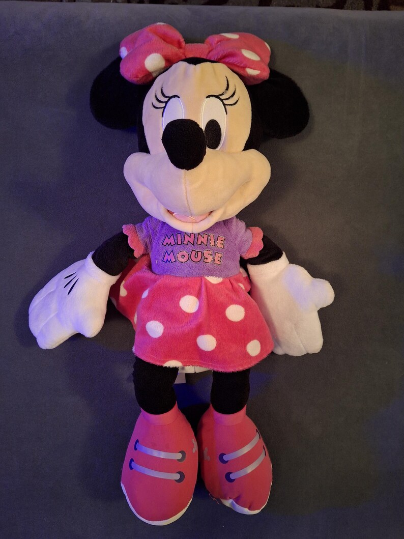 May include: A plush Minnie Mouse doll with a pink polka dot bow, dress, and shoes. The doll has black ears, a black nose, and a purple shirt with the words "Minnie Mouse". The doll has white gloves.