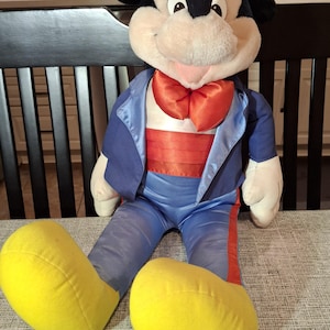 May include: A plush Mickey Mouse doll wearing a blue tuxedo jacket with a red bow tie and a red cummerbund. The doll has large yellow feet and a tag that says "applause". The doll is sitting on a gray patterned surface.