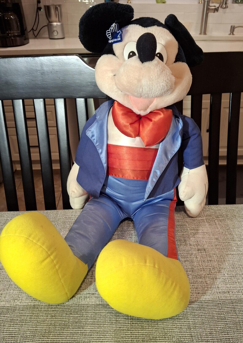 May include: A plush Mickey Mouse doll wearing a blue tuxedo jacket with a red bow tie and a red cummerbund. The doll has large yellow feet and a tag that says "applause". The doll is sitting on a gray patterned surface.