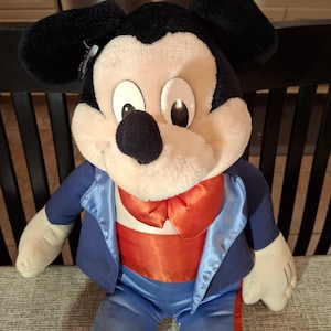 May include: A plush Mickey Mouse doll wearing a blue tuxedo jacket with satin lapels, a red bow tie, and orange cummerbund. The doll has black ears, a black nose, and large, round eyes. The doll is sitting on a gray surface.