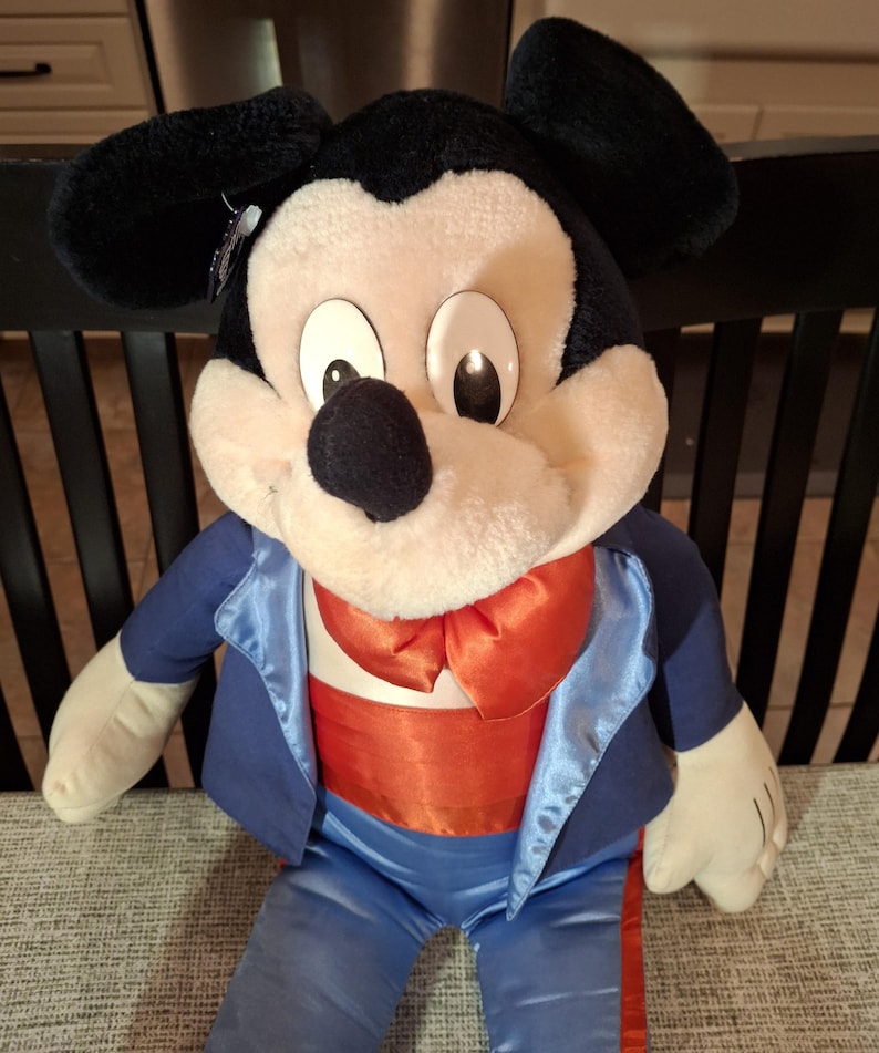 May include: A plush Mickey Mouse doll wearing a blue tuxedo jacket with satin lapels, a red bow tie, and orange cummerbund. The doll has black ears, a black nose, and large, round eyes. The doll is sitting on a gray surface.