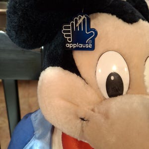May include: A plush Mickey Mouse doll with a blue and white jacket and a red bow tie. The doll has a black head and ears, and a tag that says "applause". The doll is a classic Disney character.