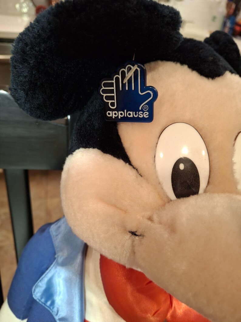 May include: A plush Mickey Mouse doll with a blue and white jacket and a red bow tie. The doll has a black head and ears, and a tag that says "applause". The doll is a classic Disney character.