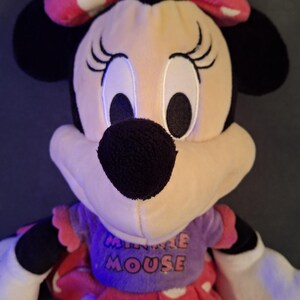 May include: A plush Minnie Mouse doll with a pink bow with white polka dots. The doll has black ears, a black nose, and large eyes. She wears a purple top with the words "MINNIE MOUSE" and a red dress with white polka dots.